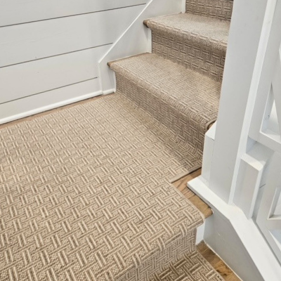 A carpeted staircase with a neutral-colored woven pattern on the treads and risers, bordered by white banisters and a horizontally paneled wall.
